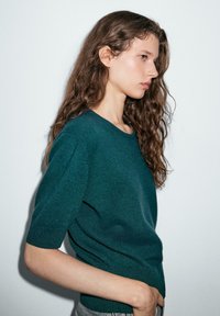 Grüner gestrickter Pullover mit kurzen Ärmeln und Rundhalsausschnitt. Glatte Textur und enganliegende Silhouette. Seitenansicht vor einem neutralen Hintergrund.