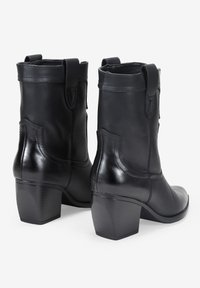 Botas de cuero negro hasta el tobillo con textura suave, que presentan un tacón apilado y costuras decorativas en los laterales.
