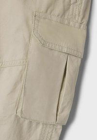 Beżowy materiał spodni cargo z dużą kieszenią z klapą, widocznymi szwami i gładką teksturą. Projekt jest funkcjonalny z praktycznymi detalami.