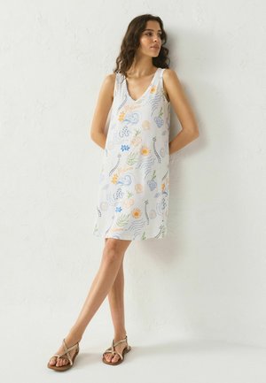 Nő ujjatlan fehér ruhában, amely színes virág- és kagylómintás, mezítláb áll egy fehér, texturált fal előtt.