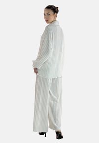 Tenue blanche texturée comprenant une chemise à manches longues et un pantalon à jambes larges avec un design plissé. Des chaussures à talons hauts noires complètent le look.