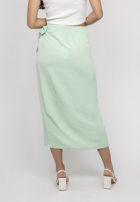 Jupe midi vert clair avec une finition texturée, dotée d'une taille élastique et d'un nœud décoratif sur le côté. Assortie à des sandales à talons bloc blanches.