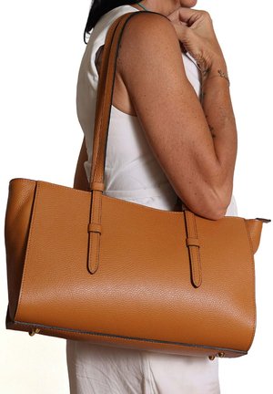 Vrouw draagt grote bruine leren shopper aan haar schouder, met een mouwloze witte top, tegen een effen witte achtergrond.