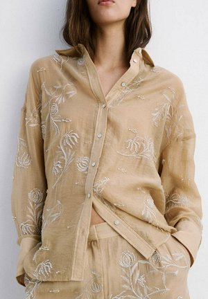 Mujer con camisa beige abotonada y pantalones a juego con bordado floral blanco, de pie con las manos en los bolsillos contra una pared blanca.