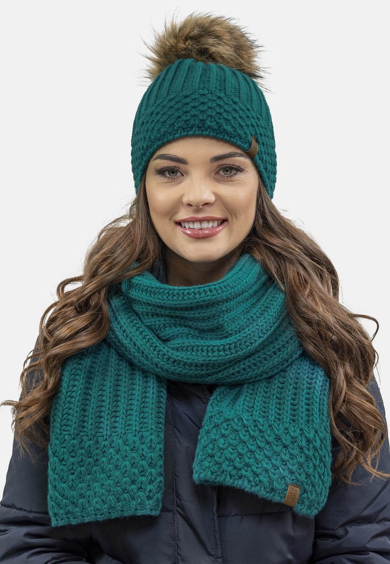 Teal strikket beanie med en pels pom-pom og matchende struktureret tørklæde. Begge har et ribbet design og læderdetaljer.