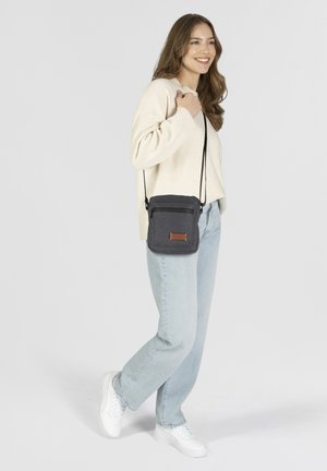 Bolso cruzado negro de tela, con un bolsillo delantero con cremallera y una etiqueta de cuero marrón, combinado con jeans de color azul claro y un suéter crema.