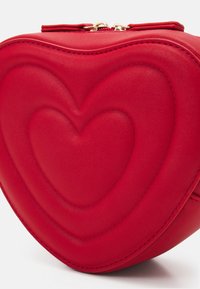 Red heart-shaped bag made of textured leather, featuring a layered heart design and gold zipper hardware. Rounded edges and stitching detail.