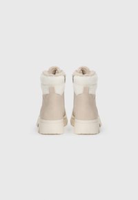Beige ankelstøvler med en fluffy hvit krage, glatt semsket skinnmateriale og en teksturert gummisåle. Har en glidelås på siden for enkel påkledning.