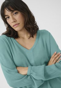 Teal, textured blouse with a V-neck and long, puffed sleeves. Fabric appears lightweight and softly draped, featuring a simple, clean design.