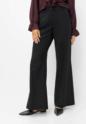 Kvinde iført sorte vidde bukser, vinrød bluse med lange ærmer og flæser samt sorte slingback sko med spids tå står på hvid baggrund.