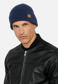Berretto in maglia a coste blu navy con una finitura texturizzata e un piccolo etichetta in pelle marrone, indossato con una giacca bomber in pelle nera.