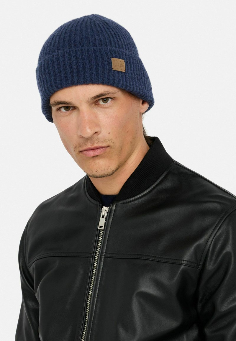 Berretto in maglia a coste blu navy con una finitura texturizzata e un piccolo etichetta in pelle marrone, indossato con una giacca bomber in pelle nera.