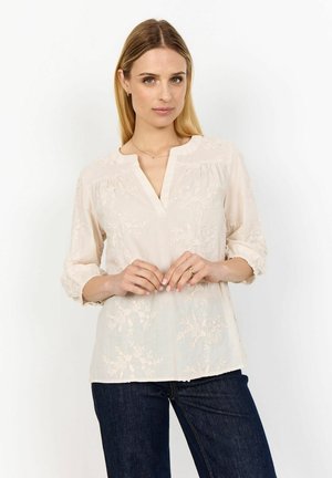 Light beige blouse with floral embroidery, V-neckline, and three-quarter sleeves, paired with dark jeans. Soft, flowing fabric texture.