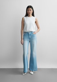 Mladá žena stojaca s jednou rukou v vrecku, oblečená v bezrukávom bielom topu, svetlomodrých vysoko pásových rozšírených džínsoch a bielych špicatých topánkach.