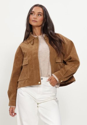 Vrouw die een bruine suède jas draagt over een lichtbeige trui en witte broek, poseert met één hand in haar zak.