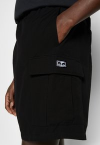 Mörkhyad person iförd svarta shorts med sidoficka och ett vitt och svart rektangulärt logo, hand delvis i fickan.