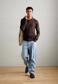 Bruine button-up cardigan, lichtblauwe oversized jeans, donkere schoenen, met een beige jas over de schouder. Soepele texturen, casual ontwerp.