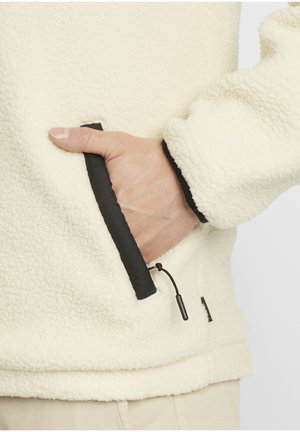 Pull-over en polaire crème avec des accents noirs. Dispose d'une poche zippée et d'un cordon de serrage. Texture douce avec une coupe décontractée.