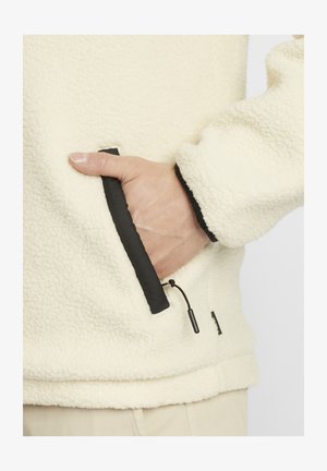 Krém színű bolyhos pulóver fekete díszítéssel. Cipzáras zsebbel és húzózsinórral rendelkezik. Puha tapintású, laza fazonú.