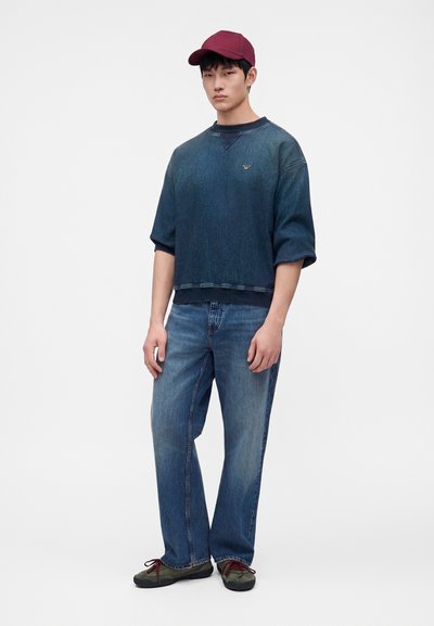 Jeune homme portant un sweat-shirt oversize bleu foncé, un jean bleu, des baskets vertes et une casquette de baseball rouge, debout devant un fond uni.