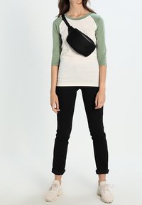 Marsupio nero con texture trapuntata indossato sopra una maglietta raglan bianca e verde, abbinato a pantaloni neri slim-fit e sneakers chiare.