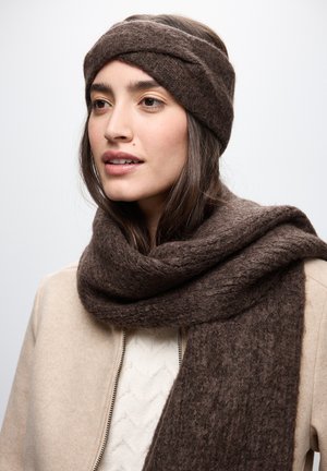 Bufanda de lana marrón tejida con una textura gruesa, drapeada alrededor del cuello; diadema marrón anudada a juego. Suéter de color crema claro debajo.