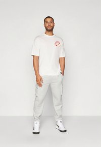 Balti medvilninė marškinėliai su raudonu grafiniu motyvu ant krūtinės, derinami su pilkomis kargo kelnėmis. Balti batai užbaigia laisvalaikio aprangą.