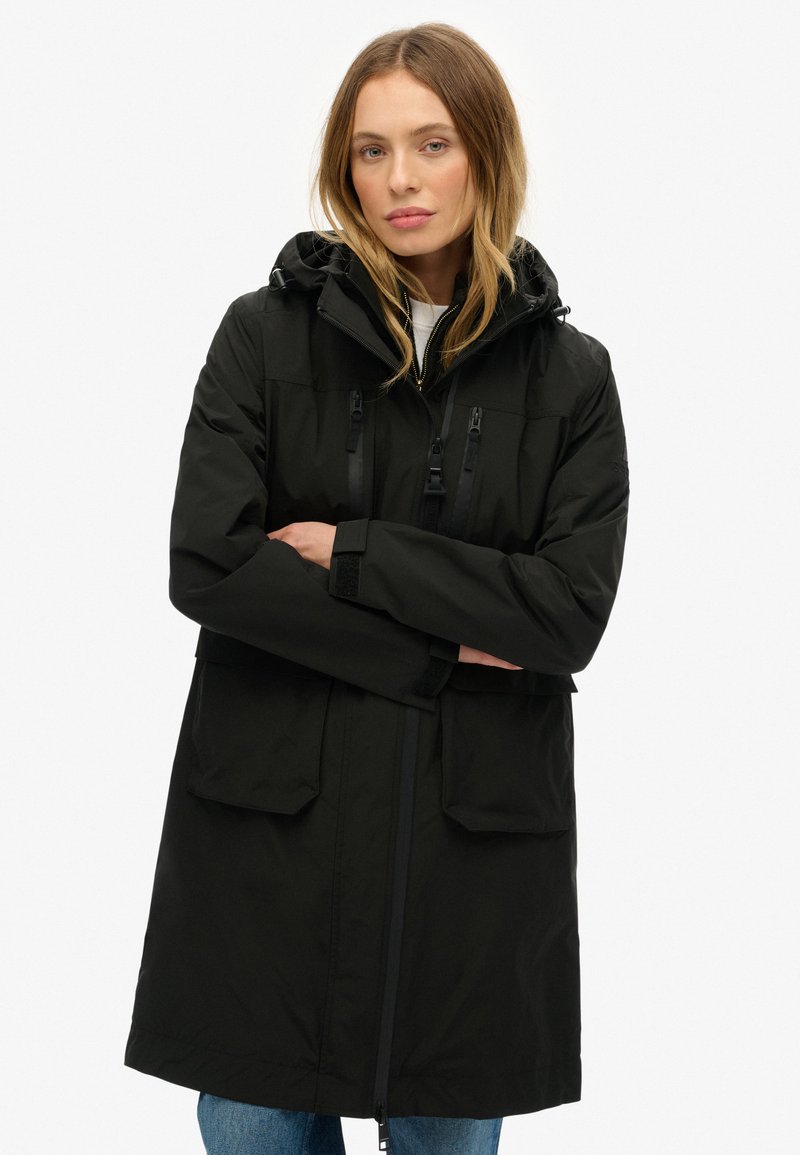 Manteau noir imperméable avec capuche, comportant deux grandes poches avant, une fermeture éclair et des cordons de serrage réglables. Tissu lisse et mat.