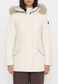 Parka color crema con cappuccio foderato in pelliccia, dotato di zip frontale, tasche laterali e una texture liscia. Il design è a maniche lunghe con una vestibilità sagomata.