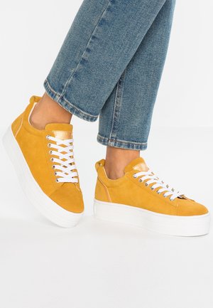 Baskets compensées en daim jaune avec des lacets blancs portées avec un jean bleu, pieds légèrement levés sur un fond blanc.