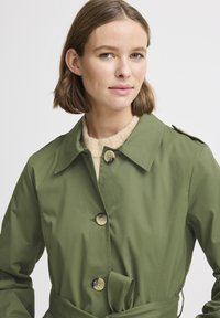 Trench coat verde oliva con colletto, chiusura a bottoni e vita con cintura. Realizzato in tessuto liscio con due grandi bottoni e tasche laterali.