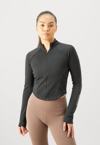 Haut de sport noir à manches longues et zippé avec une coupe ajustée. Comprend des manches raglan et des ouvertures pour les pouces. Porté avec des leggings bruns taille haute.