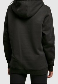 Osoba oblečená v jednoduché černé mikině a černých legínách, viděná zezadu na hladkém světlém pozadí.