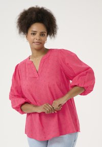 Blouse rose fuchsia avec des manches bouffantes, un col sans col et des motifs en forme de cœur texturés sur l'ensemble. Associée à un jean bleu clair.