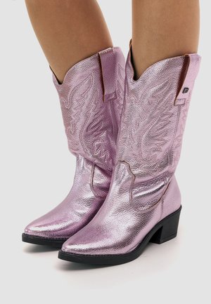 Skinnende metallisk lyserøde cowboystøvler med detaljerede syninger og sorte hæle, båret af en person, vist fra midt på læggen og ned mod en ensfarvet baggrund.