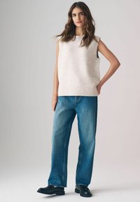Helles beige ärmelloses Stricktop mit geripptem Saum, kombiniert mit lockeren blauen Jeans und schwarzen Schuhen. Weiche Textur und lässige Passform.