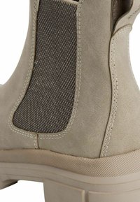Botte de cheville beige avec un panneau latéral élastique texturé gris et une semelle en caoutchouc légère. Présente des détails de couture le long des coutures.