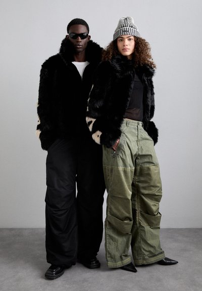 Chaqueta de piel sintética negra con acentos en beige contrastantes, combinada con pantalones cargo verde aceituna holgados. Botas de cuero negras, gorro a rayas y gafas de sol.