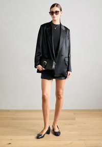 Blazer en satin noir, top noir ajusté et shorts taille haute. Sac à main noir avec une boucle dorée. Chaussures à talons blocs sombres. Sol en bois clair.