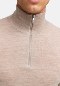 Beige kvartslång ziptröja i ribbat stickat material med hög krage och silverfärgad dragkedja. Slät textur, åtsittande design.