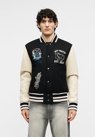 Άνδρας που στέκεται στραμμένος προς τα εμπρός φορώντας μια μαύρη και κρεμ varsity μπουφάν με γραφικά σχέδια και ανοιχτό γκρι τζιν.