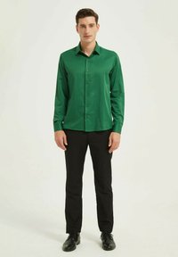 Chemise verte à manches longues en tissu lisse avec un devant boutonné, associée à un pantalon noir et des chaussures, portée par une personne se tenant debout.