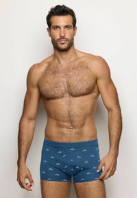 Uomo muscoloso con peli sul petto che indossa boxer blu con un motivo di piccole auto dei cartoni animati, in piedi contro uno sfondo chiaro e uniforme.