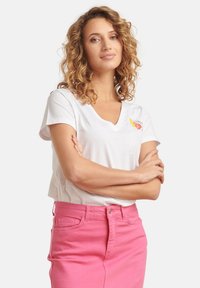 White V-neck t-shirt with a small orange and yellow graphic, paired with high-waisted pink denim skirt. Model standing with arms crossed.