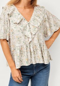 Frau in einer beigen Bluse mit Blumenmuster, fließenden Ärmeln, gerafftem V-Ausschnitt und Perlenknöpfen, kombiniert mit blauen Jeans.