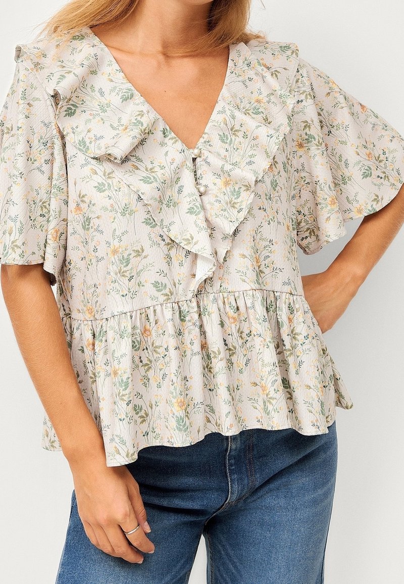 Frau in einer beigen Bluse mit Blumenmuster, fließenden Ärmeln, gerafftem V-Ausschnitt und Perlenknöpfen, kombiniert mit blauen Jeans.