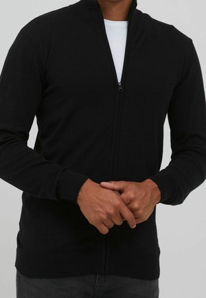 Homme portant un cardigan noir zippé sur une chemise blanche, mains lâchement jointes à la taille, sur un fond gris uni.