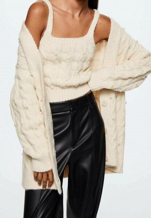 Nainen, joka käyttää kermaan vivahtavaa neulottua toppia ja siihen sopivaa ylisuurta neuletakkia helmiristikkodetaljeilla, yhdistettynä korkeavyötäröisiin mustiin nahkahousuihin.