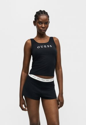 Young woman with braided hair wearing a black sleeveless GUESS tank top and black shorts with a white GUESS waistband.