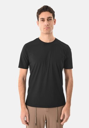 Uomo in piedi di fronte, indossa una t-shirt nera a maniche corte e pantaloni color sabbia con coulisse, su sfondo bianco.